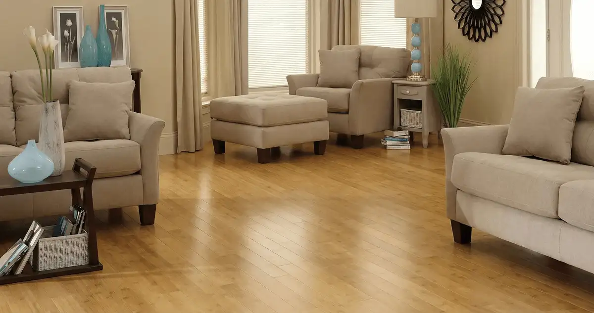 A living room with professionally installed bamboo flooring, beige sofas, a matching ottoman, a side table with a blue lamp, vases, framed pictures, and large windows with blinds and curtains in Bakersfield, CA