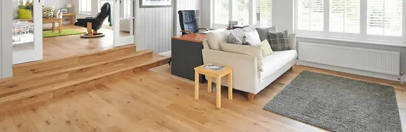 A modern living room with professionally installed laminate flooring, a beige sofa, a small side table, a gray rug, and large windows with white shutters in Bakersfield, CA